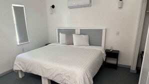 A bedroom with a large bed, a bedside table, a wall-mounted air conditioner, and a window.