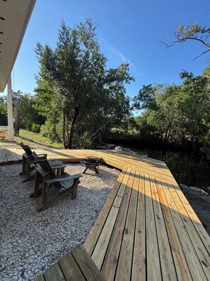 Eine Holzterrasse mit Sitzgelegenheiten und Grill, umgeben von Bäumen und einem Gewässer.