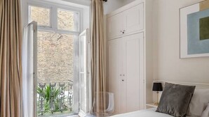 A bedroom with a bed, a nightstand, a lamp, a window with curtains, and a view of a brick wall and greenery outside. - 1 Bed Flat in Earl\'s Court (Greater London)