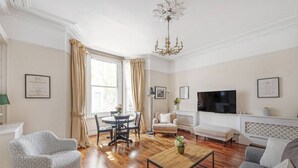 A living room with a wooden floor, a sofa, a coffee table, a TV, and a chandelier. - 1 Bed Flat in Earl\'s Court (Greater London)