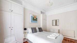A hotel room with a large bed, white wardrobe, bedside table, and a clear chair. - 1 Bed Flat in Earl\'s Court (Greater London)