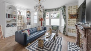 A living room with a blue sofa, a coffee table, and a fireplace. There are bookshelves and a chandelier. - Blossom House New 3Bed house in Barons Court (Greater London)