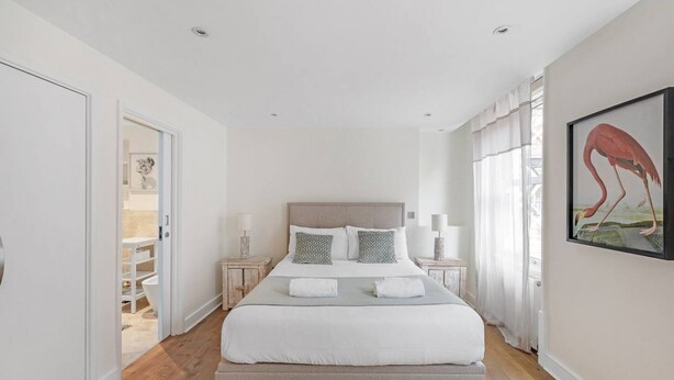 A bedroom with a large bed, bedside tables, a nightstand, a framed picture on the wall, and a door leading to a bathroom. - New 2Bed with Air Con in Kensington (London)