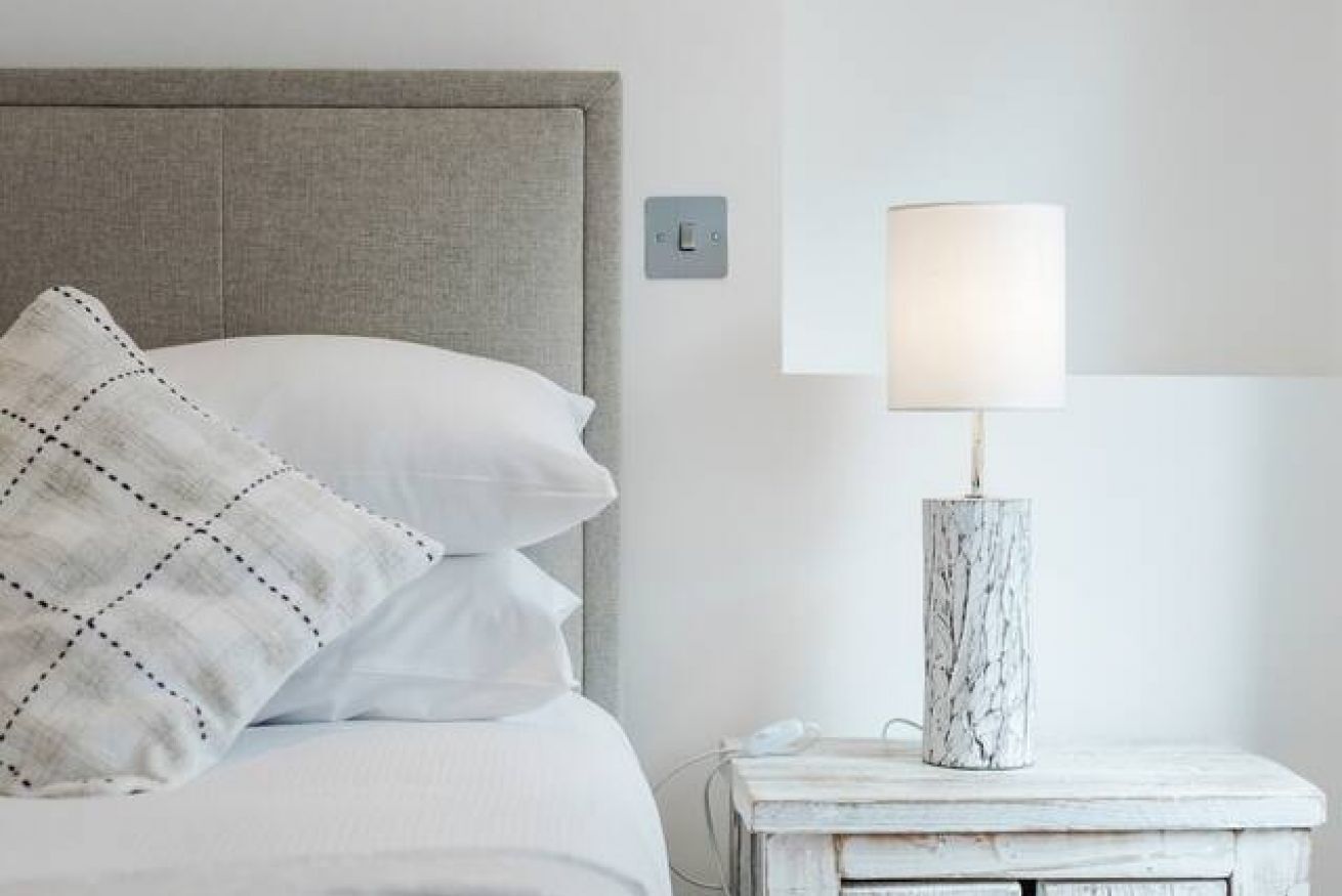 Una cama bien hecha con un cabecero gris, una almohada blanca con diseño geométrico y una lámpara blanca sobre una mesita de noche blanca.