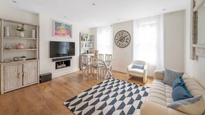 A living room with a TV, wooden cabinets, a dining table with chairs, a sofa, and a patterned rug. - New 2Bed with Air Con in Kensington (London)
