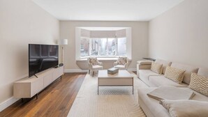 A modern living room with a flat-screen TV, a sofa, a coffee table, and a window with a view of buildings. - New 3Bed in Knightsbridge (Greater London)