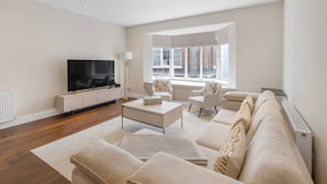 A modern living room with a flat-screen TV, a sofa set, a coffee table, and a large window offering a view of the street. - New 3Bed in Knightsbridge (Greater London)