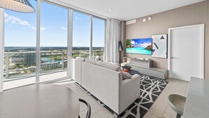 A modern living room with a large window, a flat-screen TV, and a dining area. - 2BR Ocean Penthouse: Private Terrace & Hot Tub (Hollywood)