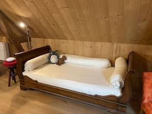 Una cama de madera en una habitación con techo inclinado, una mesita de noche con lámpara y un taburete rojo.