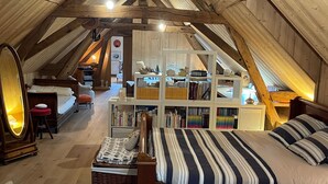 Une chambre mansardée avec un plafond en pente en bois, un grand lit avec une literie à rayures, une bibliothèque et un petit tabouret.