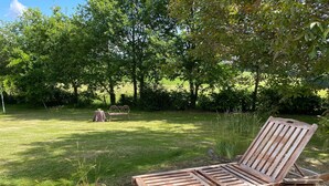 Un fauteuil de jardin en bois muni de roulettes, un bac à fleurs en pierre orné de plantes, et un banc en métal, le tout installé dans un espace extérieur verdoyant, avec des arbres en arrière-plan.
