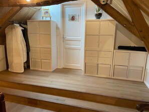 Una habitación con techo inclinado, vigas de madera, un armario blanco con varios cajones, un perchero con una prenda blanca y una puerta con un pequeño marco de fotos.