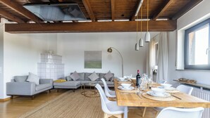 Ein modernes Wohnzimmer mit Essbereich, mit Holzboden, einem grauen Sofa, einem Esstisch mit weißen Stühlen und einem großen Fenster.