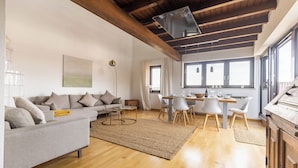 Ein modernes Wohnzimmer mit einer Couch, einem Esstisch und Stühlen. Es gibt eine Holzbalkendecke und große Fenster.