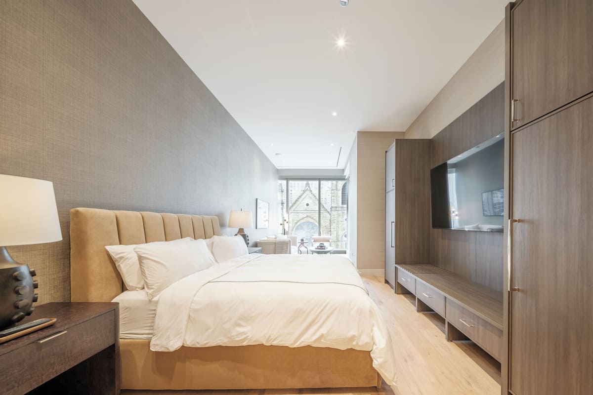 Ein modernes Schlafzimmer mit einem großen Bett, einem Fernseher und Blick auf ein Gebäude durch das Fenster.