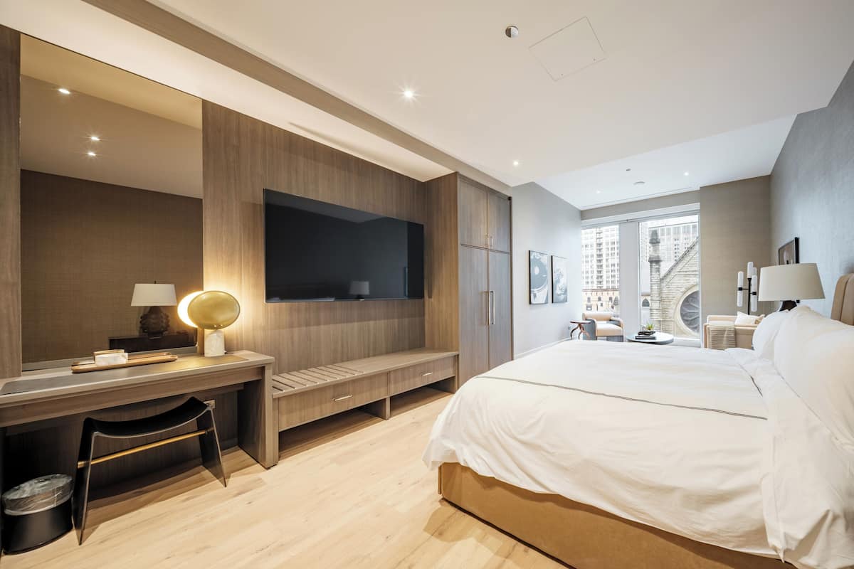 Ein modernes Hotelzimmer mit einem großen Bett, einem Schreibtisch mit Lampe, einem Fernseher und Blick auf die Stadt durch ein großes Fenster.