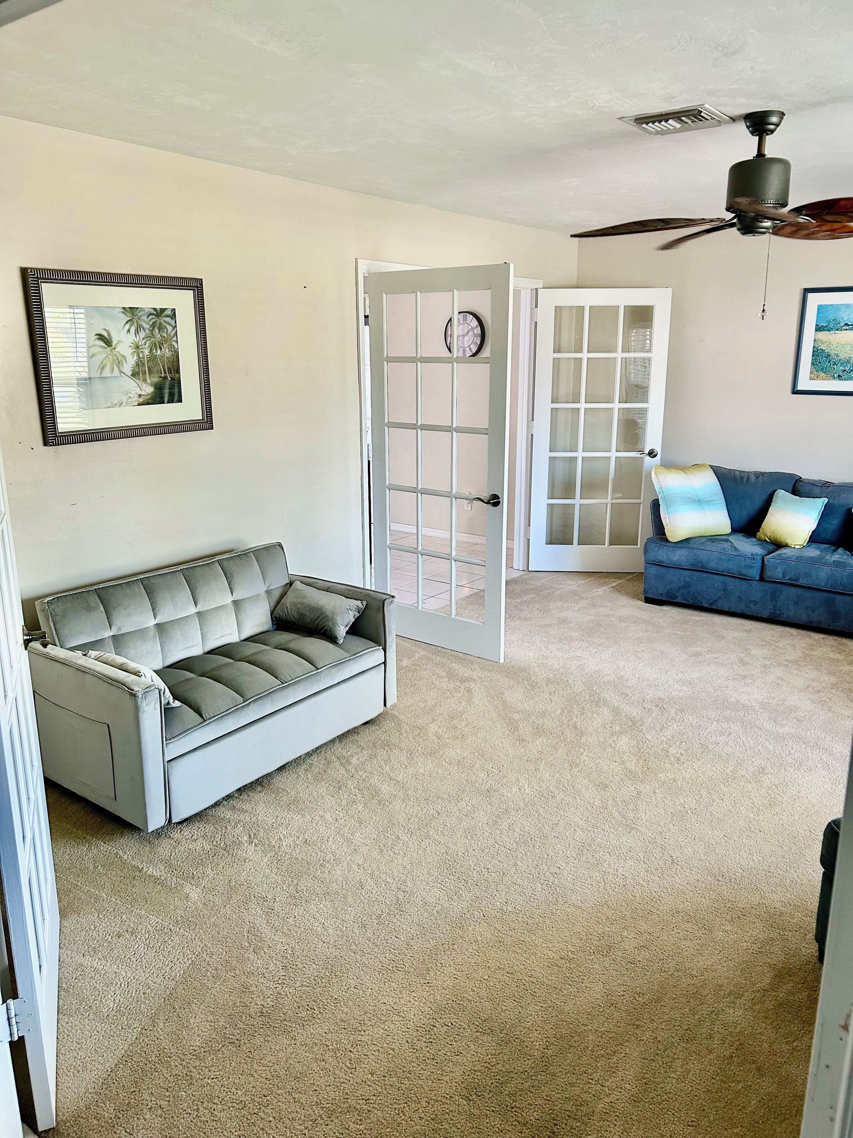 A living room with a grey sofa, a blue sofa with cushions, a ceiling fan, and a door leading to another room.