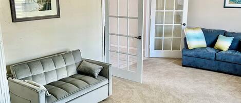 A living room with a grey sofa, a blue sofa with cushions, a ceiling fan, and a door leading to another room.