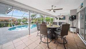 A covered outdoor dining area with a pool view, wicker chairs, a ceiling fan, and a TV mounted on the wall. - 3-bedroom & den house in welcoming Cape Coral with Pool, centrally located!🌴 (Cape Coral)