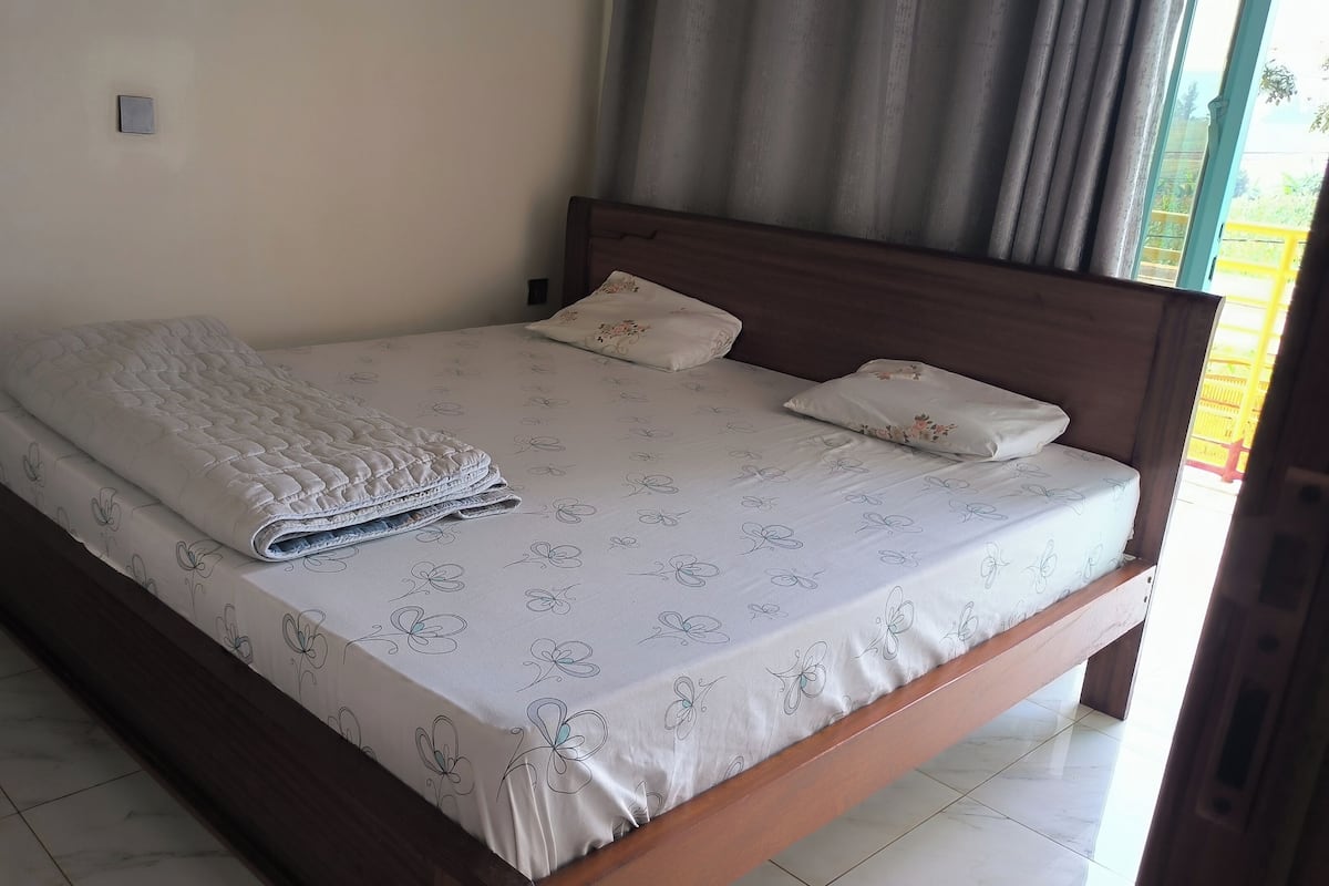 A neatly made bed with a floral-patterned bedspread and pillows, a wooden headboard, and a tiled floor.