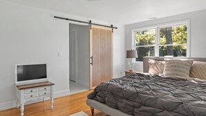 A bedroom with a bed, a TV on a nightstand, a sliding door, and a window with a view of trees. - Cozy North Fork Home Close to Beaches, Wineries, and Town, Perfect for Families (East Marion)