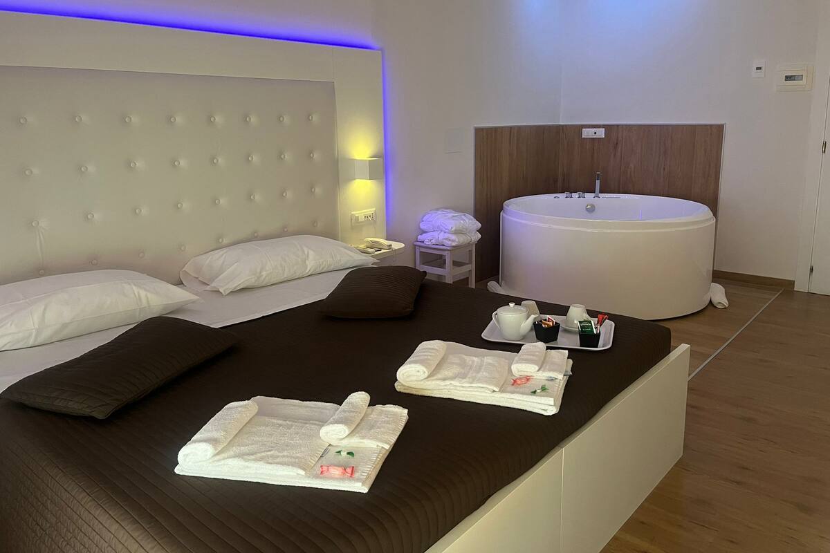 Hotelkamer met een groot bed, nachtkastje met telefoon en lamp, badkuip, en handdoeken en toiletartikelen op het bed.