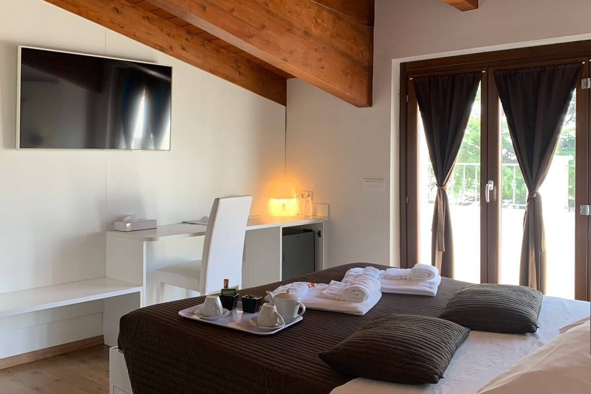 Hotelkamer met een houten plafond, een bed met een dienblad vol witte kopjes en een theeset, een bureau met stoel, en een aan de muur bevestigde televisie.
