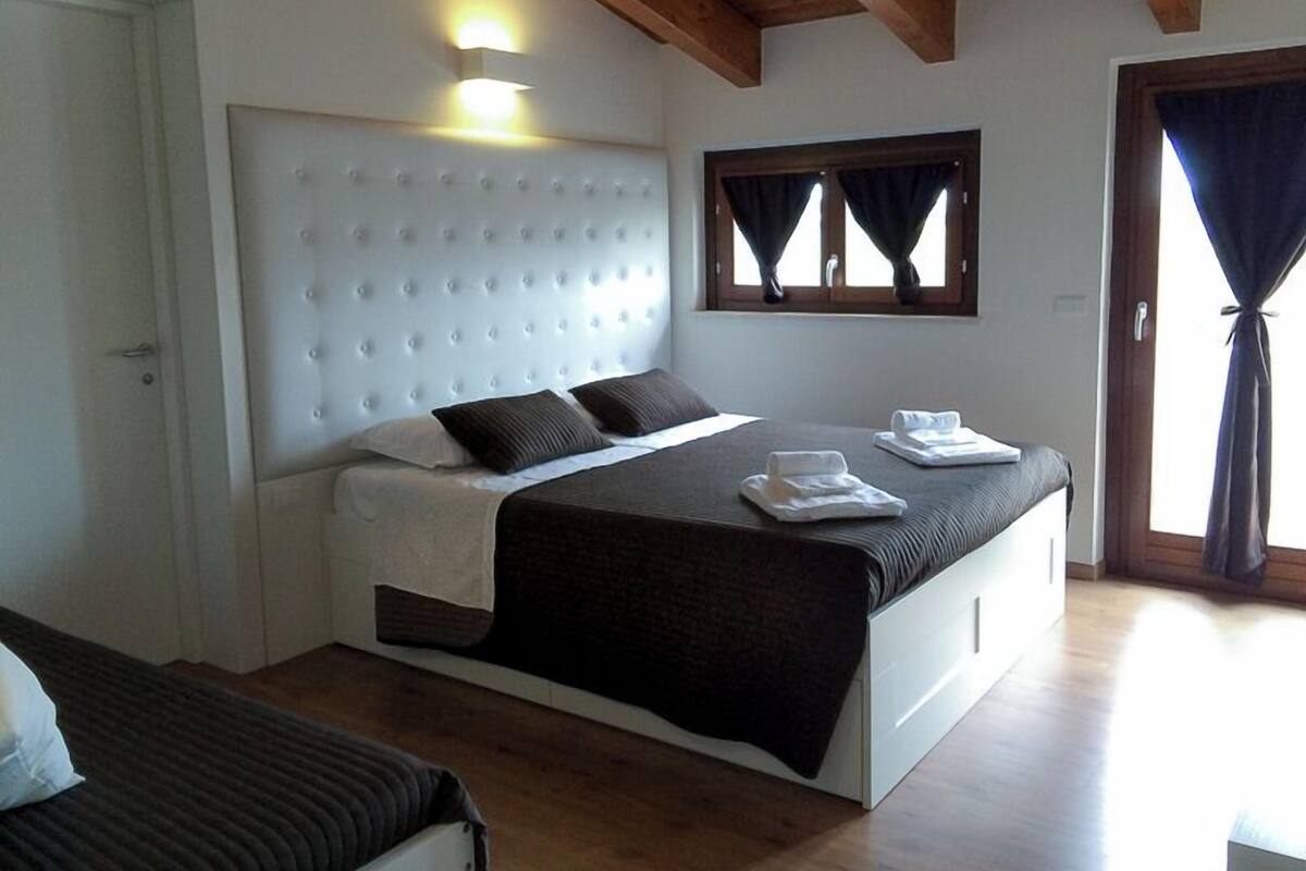 Een slaapkamer met een gestoffeerd hoofdeinde, een bed met donkere beddengoed, een nachtkastje en een raam met gordijnen.