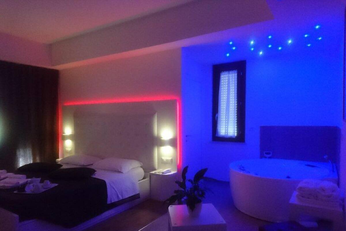 Een moderne hotelkamer met een bed, nachtkastjes, een klein tafeltje met een plant, een badkuip en een raam met zonwering.
