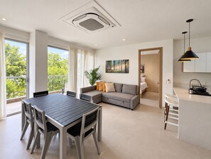 A modern living space with a dining area, a sofa, a kitchen, and a large window with a view of greenery. - Baia Kristal Prive | 2 Bedrooms | Sleeps 8 | P31L (Cartagena de Indias)