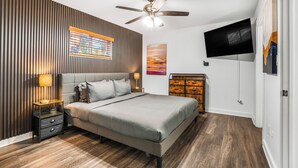 Un dormitorio con una cama, ventilador de techo, televisión, mesita de noche, lámpara y cómoda.