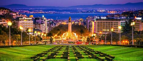 Un paisaje urbano nocturno con una estatua destacada en el centro, rodeado de edificios bien iluminados y un parque con parterres de césped geométricos.