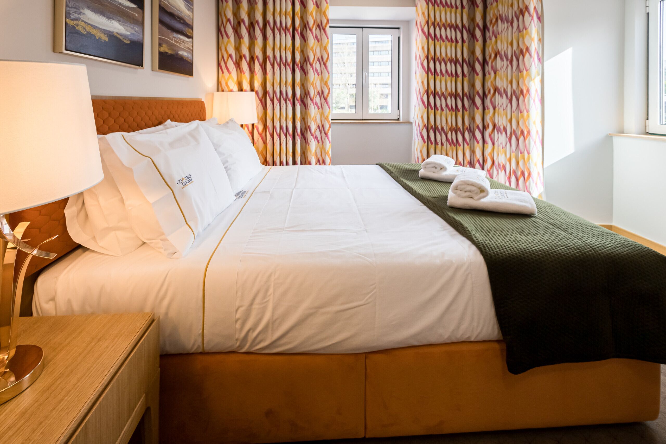 A hotel room with a large bed, a wooden headboard, a bedside table with a lamp, and colorful curtains.