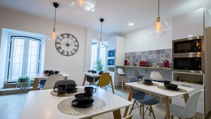 Una cocina moderna con armarios blancos, un microondas y una estufa. Hay dos mesas de comedor con sillas, y un reloj de pared grande.