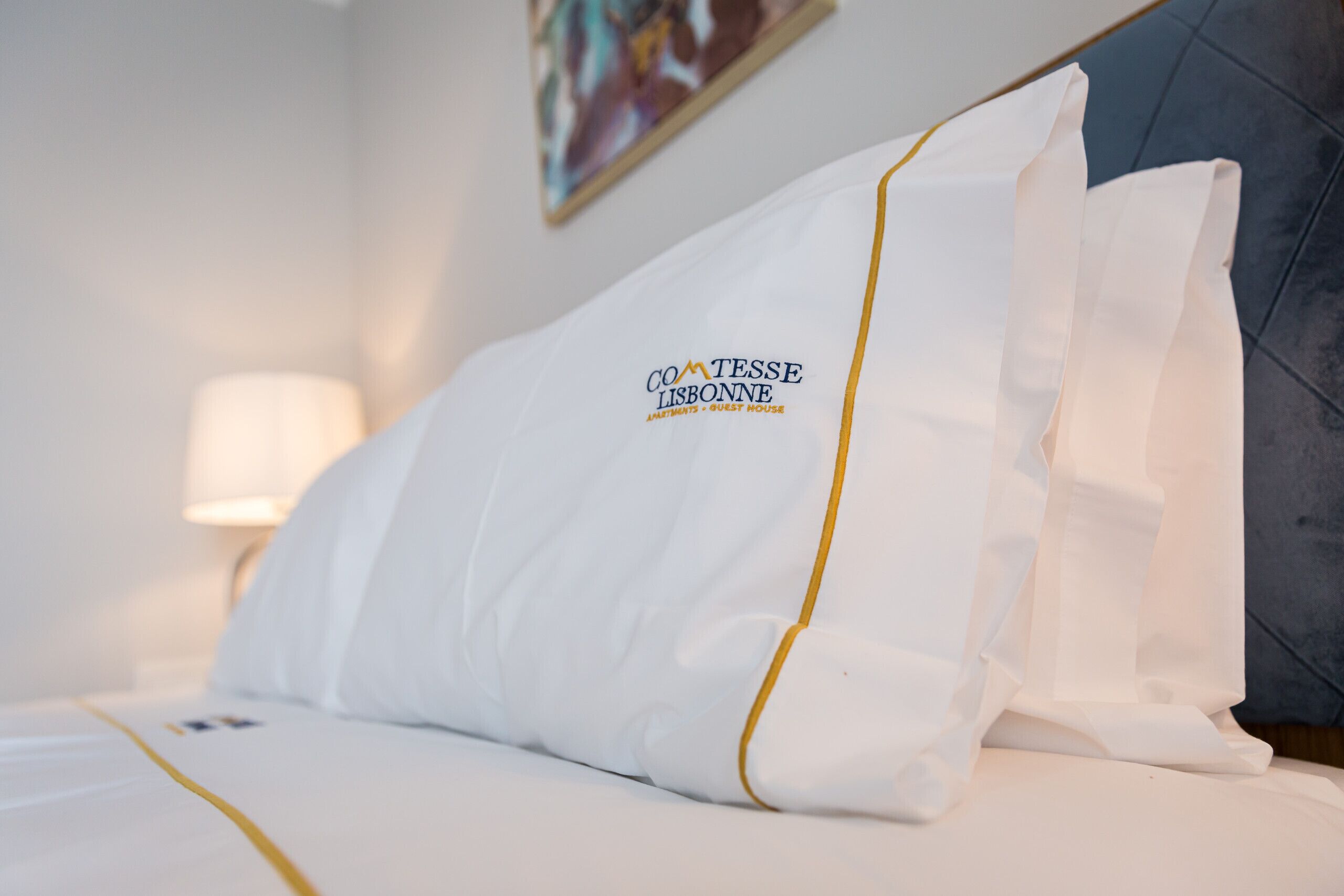 En hotellsäng med vita sängkläder och en kudde prydd med logotypen ”Comtesse Lisbonne”.
