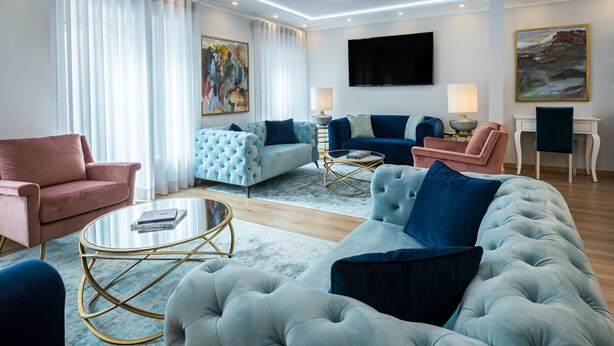 Una sala de estar moderna con un sofá grande de color gris, un sillón rosa y un sillón azul. Hay un televisor de pantalla plana montado en la pared, una mesita con una lámpara, y un cuadro en la pared.
