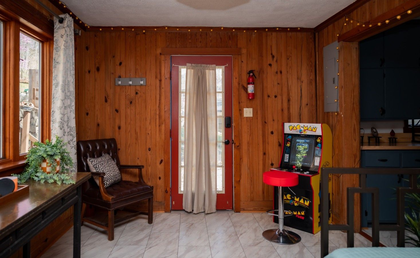 Une pièce aux murs en bois, une porte ornée de rideaux, un fauteuil en cuir, une petite table avec une plante et une borne d’arcade Pac-Man.