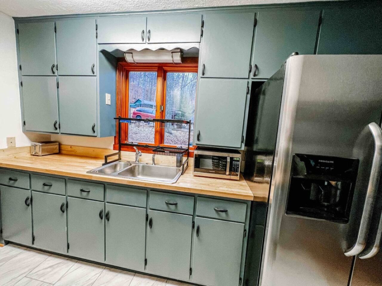 A kitchen with grey cabinets, a stainless steel refrigerator, and a window with a view of the outside.