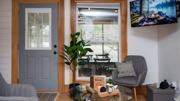 Una habitación con una puerta gris, un ventanal que da al exterior, un sillón gris, una mesa de centro de vidrio y un televisor fijado a la pared.