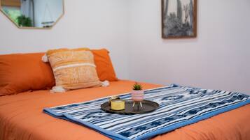 Un lit avec une literie orange, une couverture rayée bleue et blanche, une petite plante en pot et un plateau avec une bougie et un récipient.