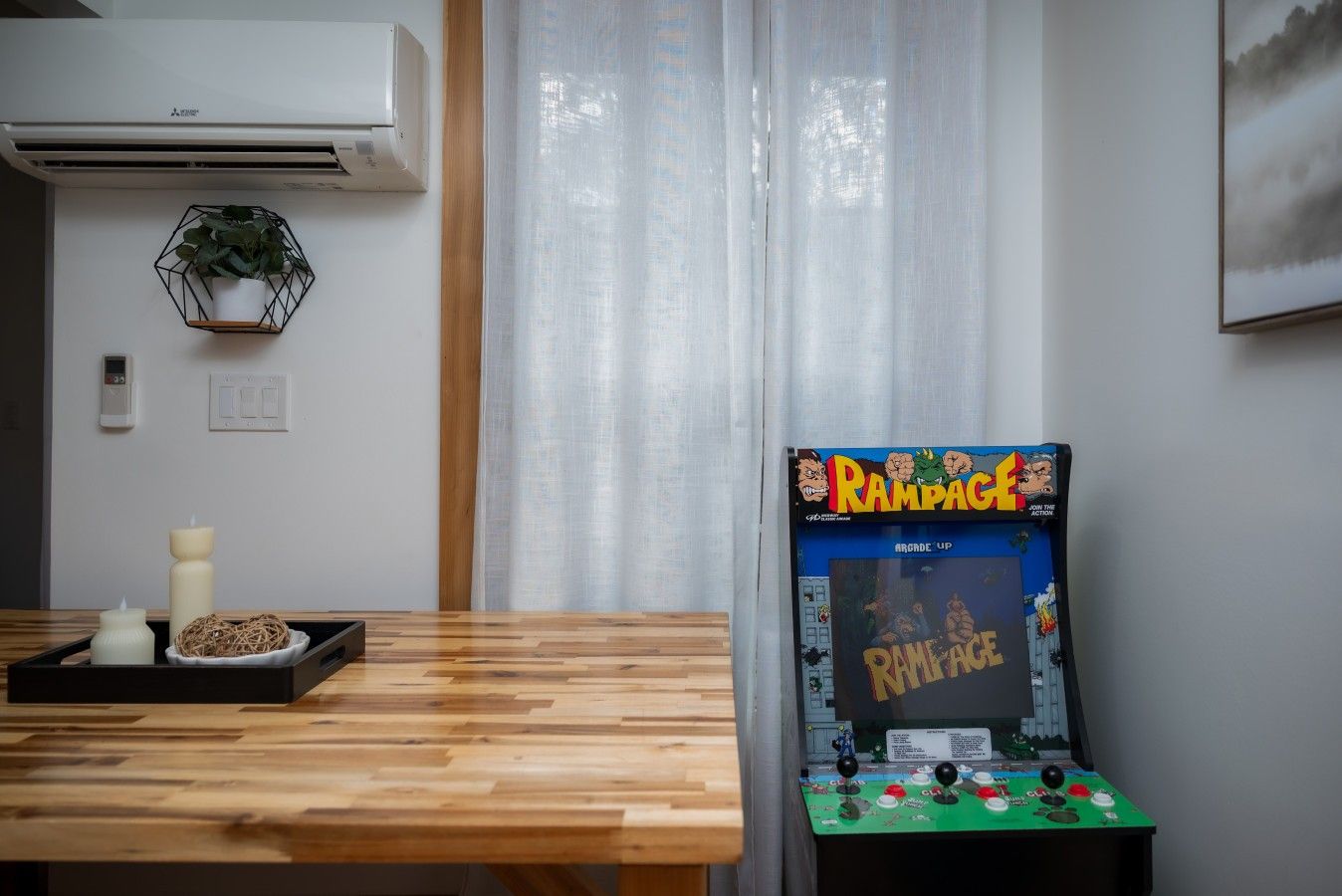 A room with a wooden table, a wall-mounted air conditioner, a wall-mounted thermostat, a wall-mounted light switch, a wall-mounted electrical outlet, a wall-mounted decorative item, a wall-mounted artwork, and a video game console.