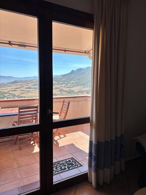 Una habitación con puerta de vidrio corredera que da a un balcón exterior. El balcón tiene una silla de madera y una mesita.