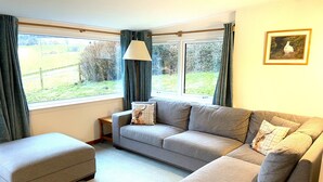 A living room with a grey sofa, an ottoman, a small table, and a lamp. There is a window with curtains and a framed picture on the wall. - The Mill - Amazing spacious mountain escape in Speyside & the Cairngorms (Ballindalloch)