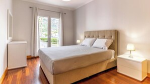 A bedroom with a bed, bedside tables, a lamp, a radiator, and a view of greenery outside. - Ocean View Flat (Câmara de Lobos)