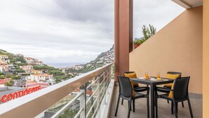 A balcony with a table and chairs, overlooking a hillside town. - Ocean View Flat (Câmara de Lobos)