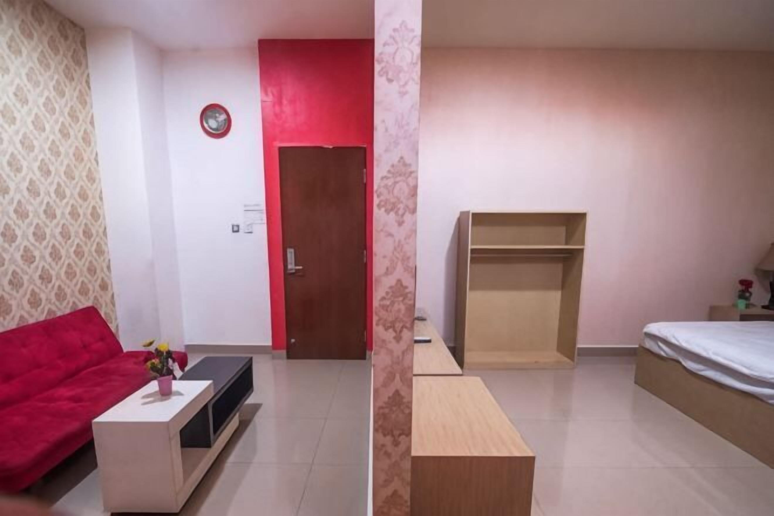 Um quarto com um sofá vermelho, uma mesa pequena com um vaso, um banco de madeira, uma cama e uma porta.