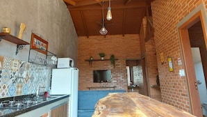A kitchen with a brick wall, wooden ceiling, and a countertop with a stove. There is a television mounted on the wall and a window with blinds. - Villa da Serra chalé (Lima Duarte)