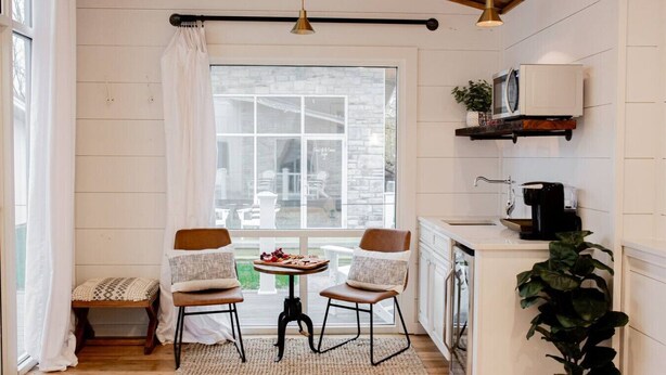Una cocina acogedora con dos sillas, una mesita con aperitivos, una cafetera y vistas a través de un ventanal grande.