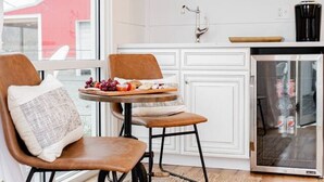 Una cocina moderna con techo de madera, un comedor con dos sillas, una mesa pequeña con una bandeja de comida y un ventanal con vista a un edificio rojo.