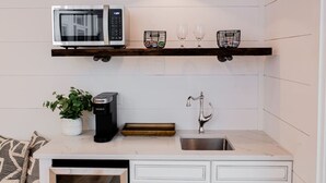 A kitchen with a microwave, coffee maker, and sink. There are two shelves above the sink with glasses and a basket.